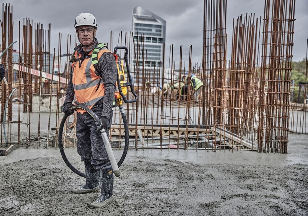 Person wearing a safety helmet and high-visibility vest, operating a DEWALT concrete vibrator tool (DCPS310N) at a construction site with rebar structures and wet concrete.