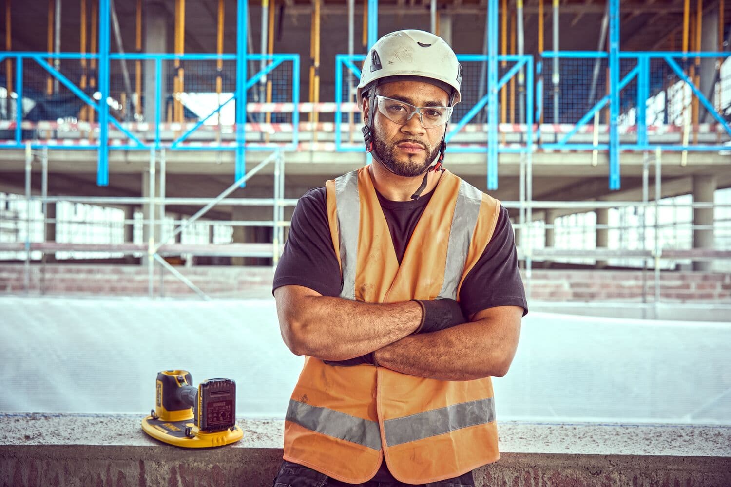 A DEWALT DCE590 tool and battery charger placed on a concrete ledge in front of a construction worker wearing an orange reflective safety vest and helmet at a building site with scaffolding and blue railings in the background.