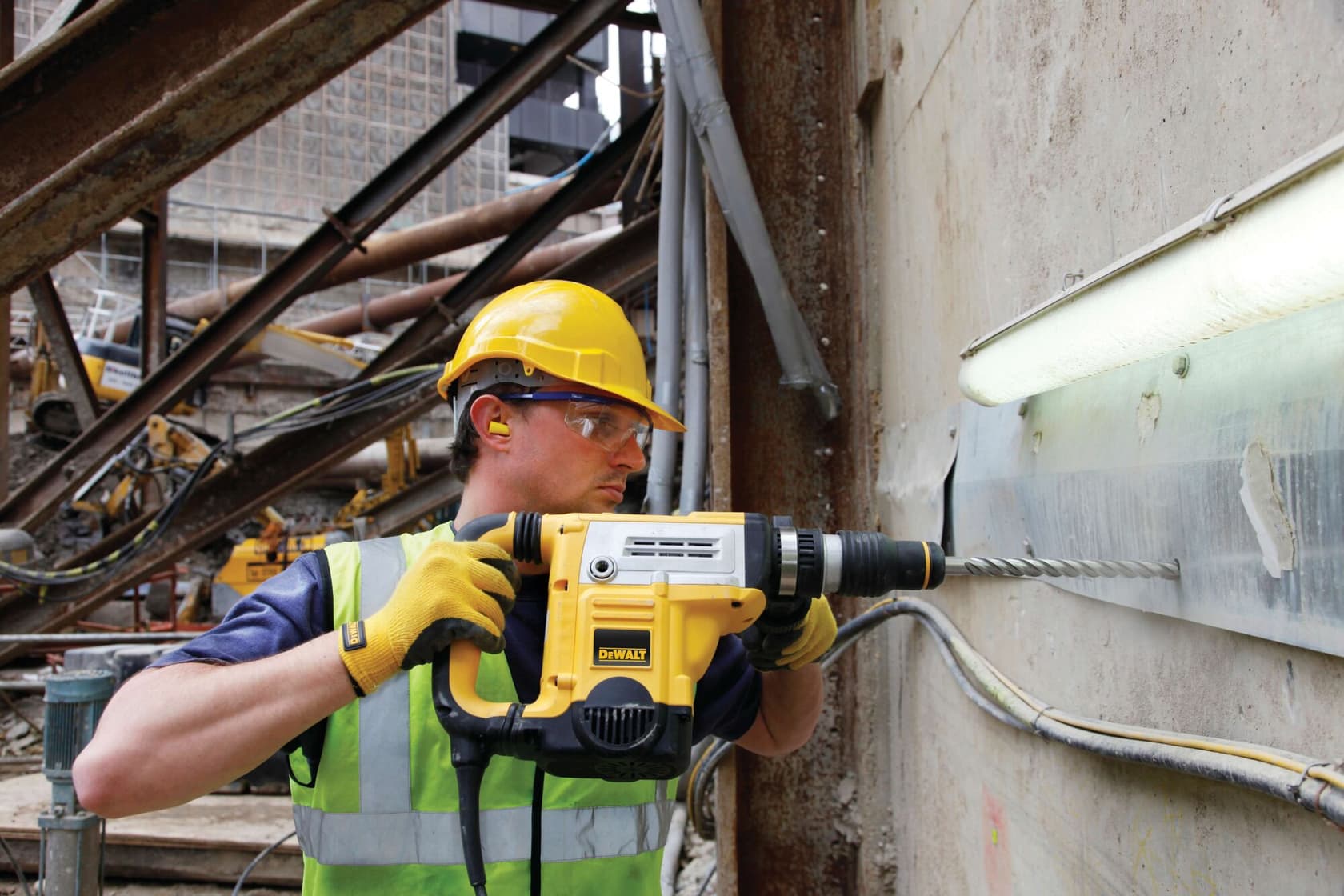 A person wearing safety gear is using a DEWALT rotary hammer drill (SKU: DT60844) to drill into a concrete wall at an