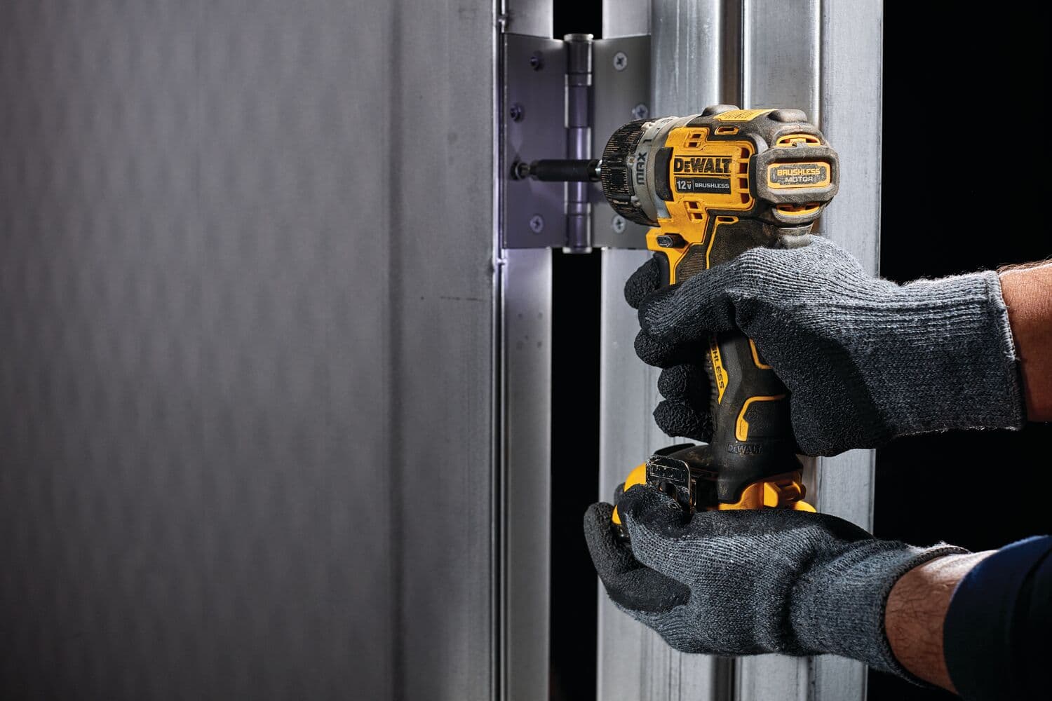 A person wearing grey gloves is using a yellow DEWALT cordless drill to install or tighten screws on a metal hinge attached to a metallic door frame.