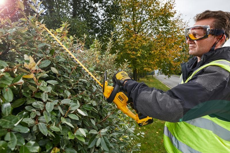 A person wearing safety gear and gloves is using a DEWALT DCM563PB cordless hedge trimmer to cut a dense bush outdoors. The person's face is blurred for privacy.
