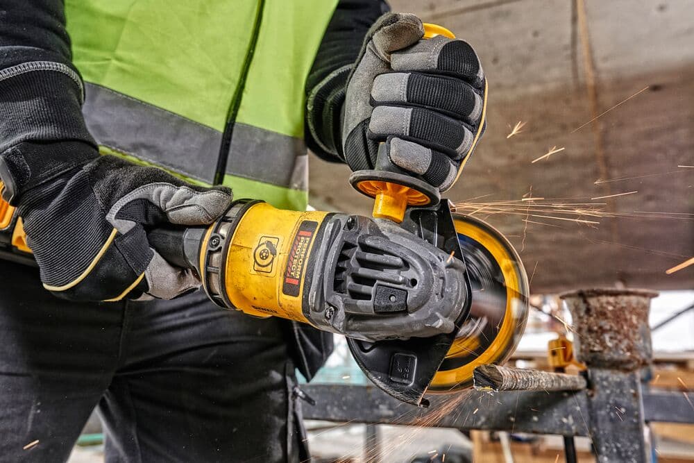 A person wearing safety gloves and a high-visibility vest is using a yellow DEWALT angle grinder to cut through a metal bar