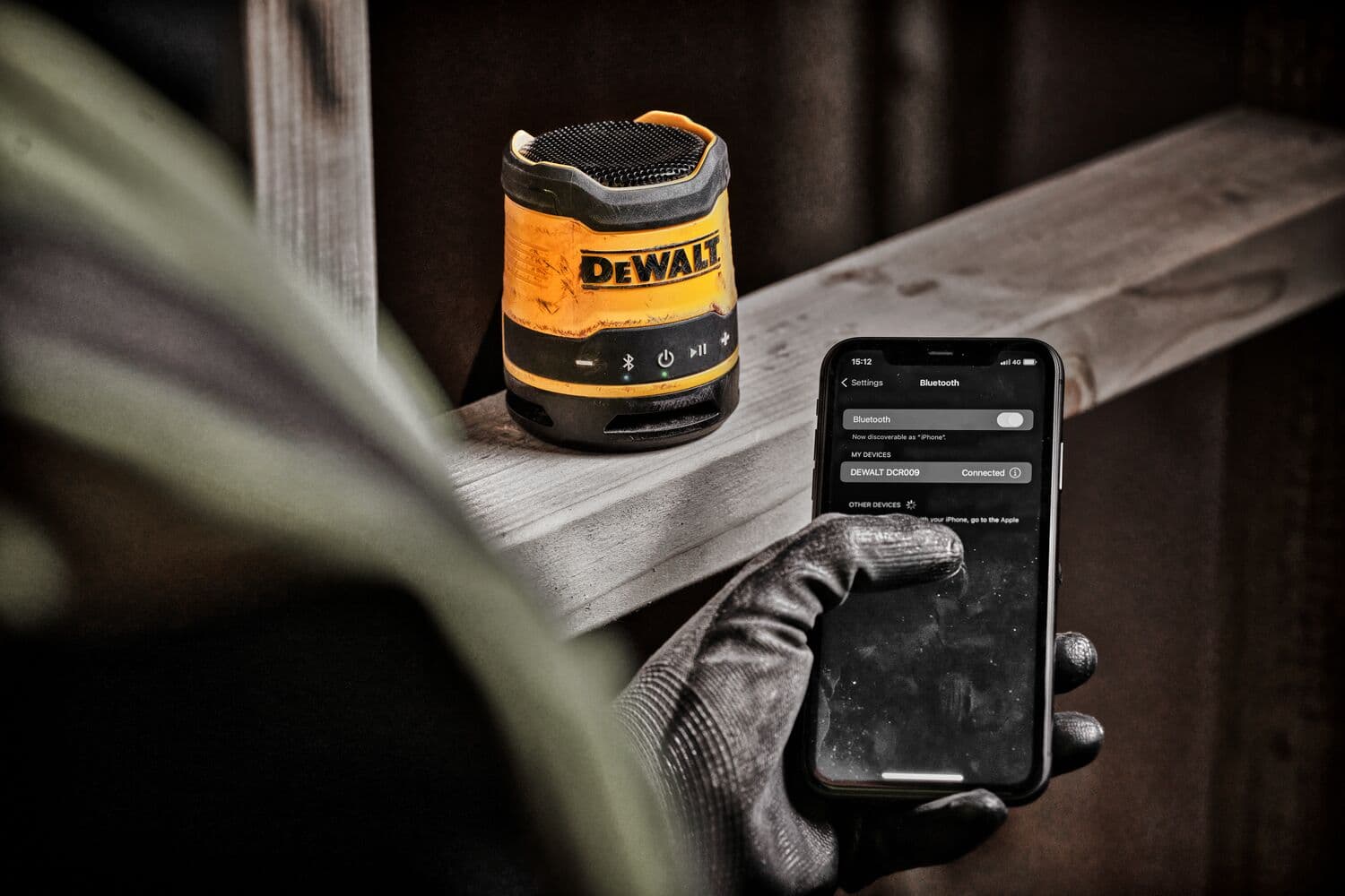 A DEWALT DCR009 portable Bluetooth speaker sits on a wooden surface, while a person wearing a work glove operates a smartphone showing the speaker connected via Bluetooth.