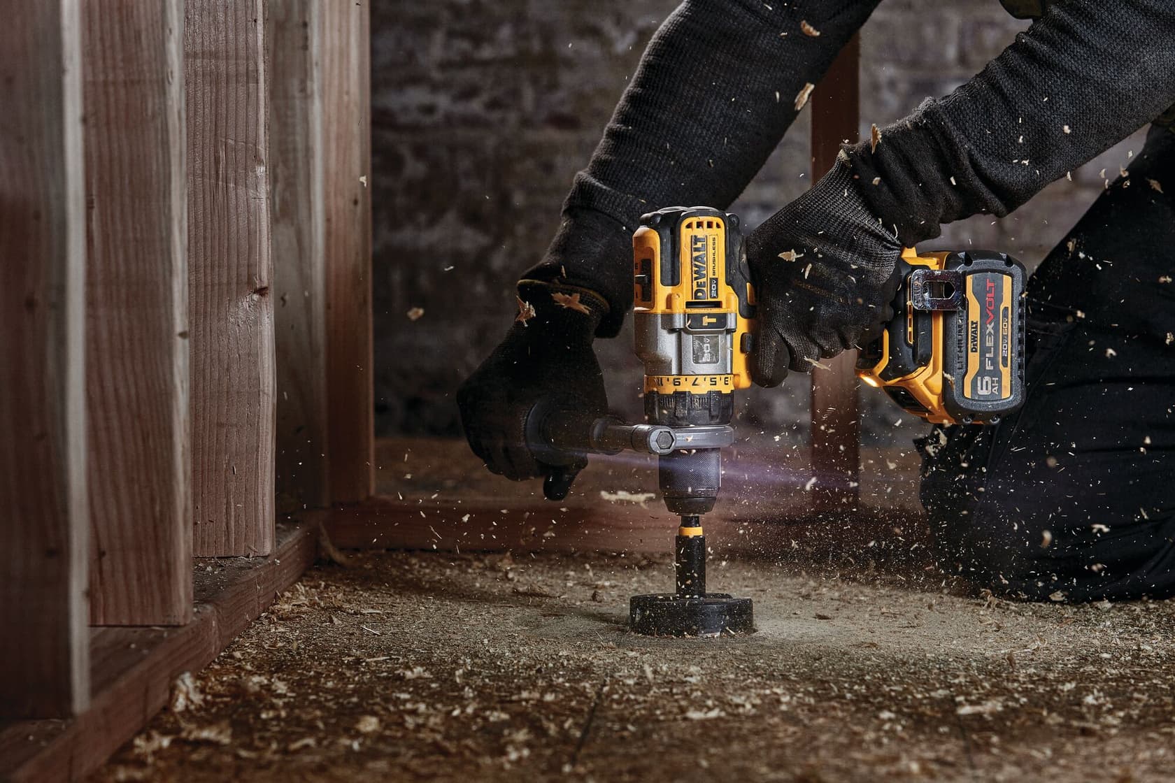 A person uses a DEWALT cordless power drill, model DCD999T1, to drill into a wooden floor. The person is wearing black gloves and working in a construction or renovation setting, with wood shavings flying around.