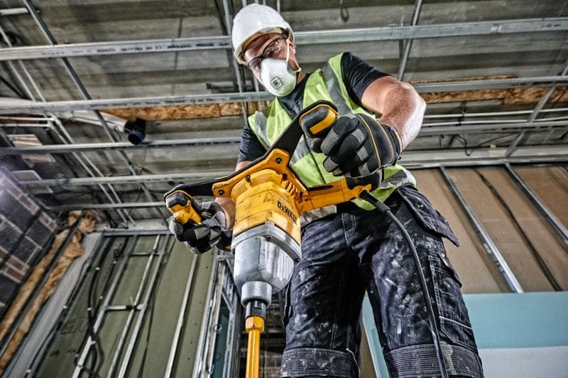 A construction worker wearing a safety vest, gloves, and a helmet is operating a DEWALT DWD241 electric mixer drill inside a