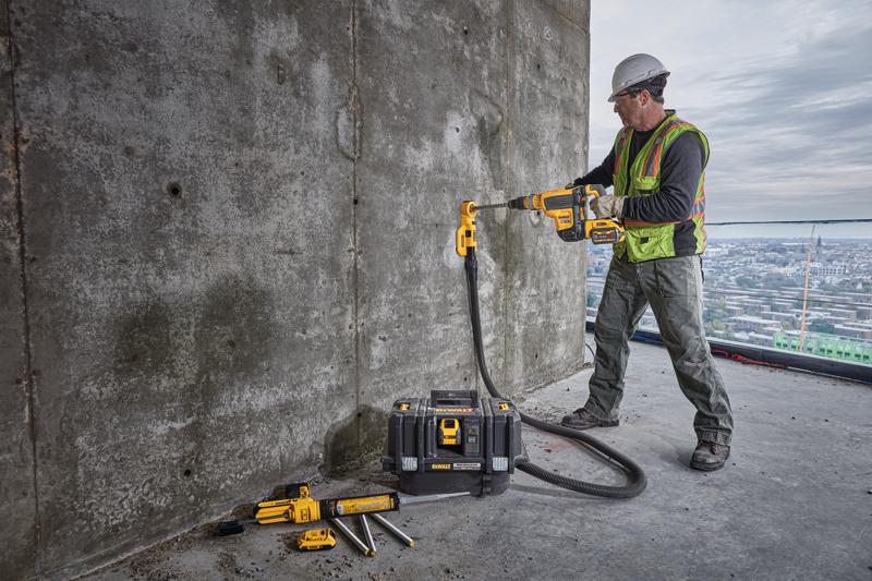 A construction worker wearing a hard hat and safety vest uses a DEWALT power tool to drill into a concrete wall. The worker is outdoors at a construction site, with a DEWALT dust extraction attachment connected to the drill by a hose and a DEWALT tool box and accessories placed nearby on the ground.