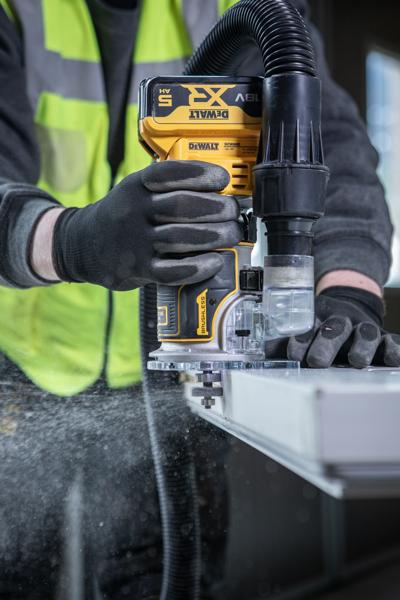 A person wearing gloves and a high-visibility vest is using a DEWALT cordless router on the edge of a work surface, with a dust extraction hose attached.