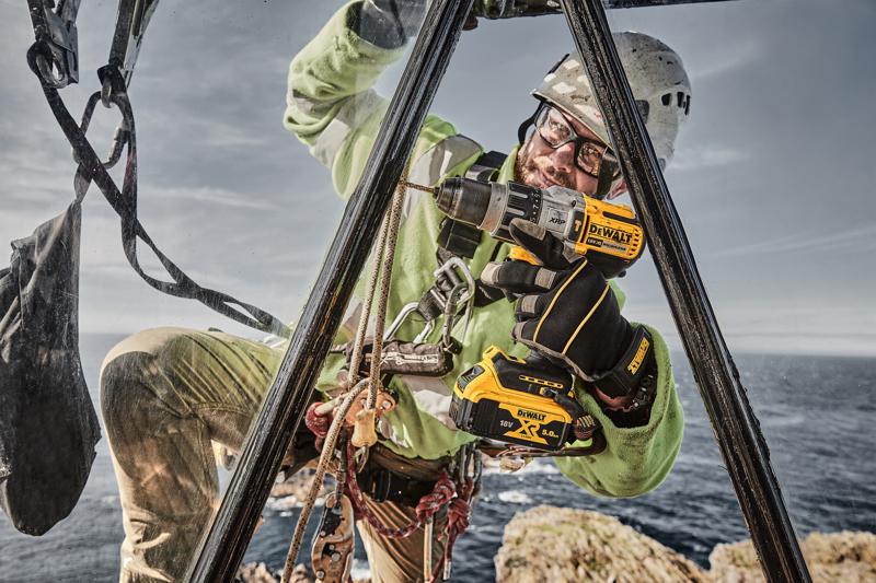 A worker wearing safety gear, including gloves and a helmet, uses a DEWALT DCD996P2 cordless drill at a high outdoor location near the sea, with ropes and harness visible for safety.