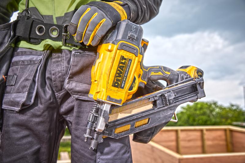 Close-up of a person wearing work gloves holding a DEWALT DCN930N nailer at a construction site, with a wooden structure in the background.