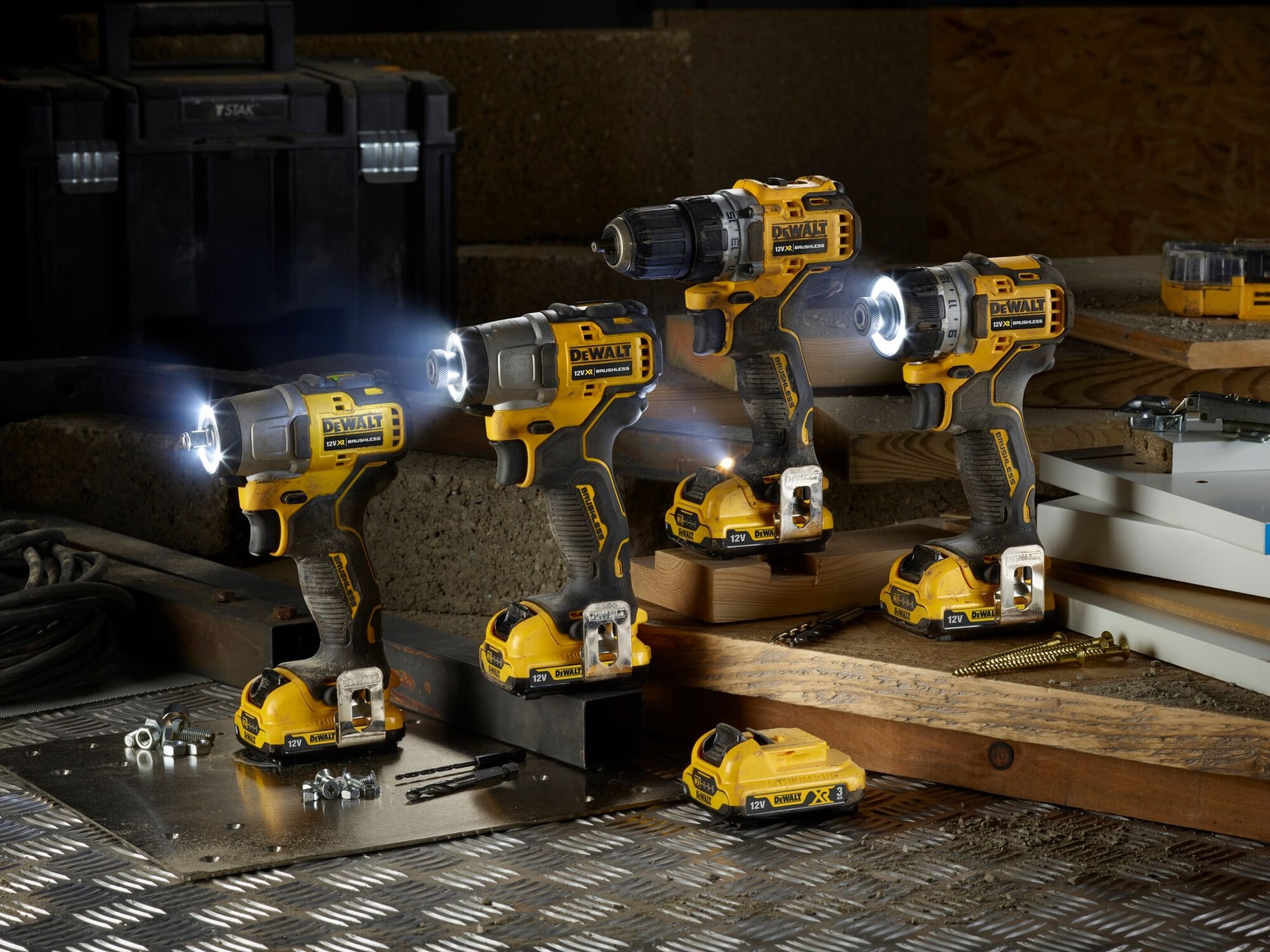 Three DEWALT cordless power tools with LED lights are displayed on a workbench along with a DEWALT battery, metal parts, drill bits, screws, and other construction materials. The background includes a black toolbox and various wooden surfaces.
