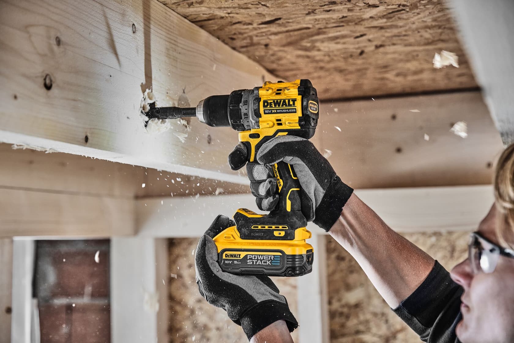 A person wearing gloves uses a DEWALT DCD800H2T cordless drill to make a hole in a wooden beam, with wood shavings flying