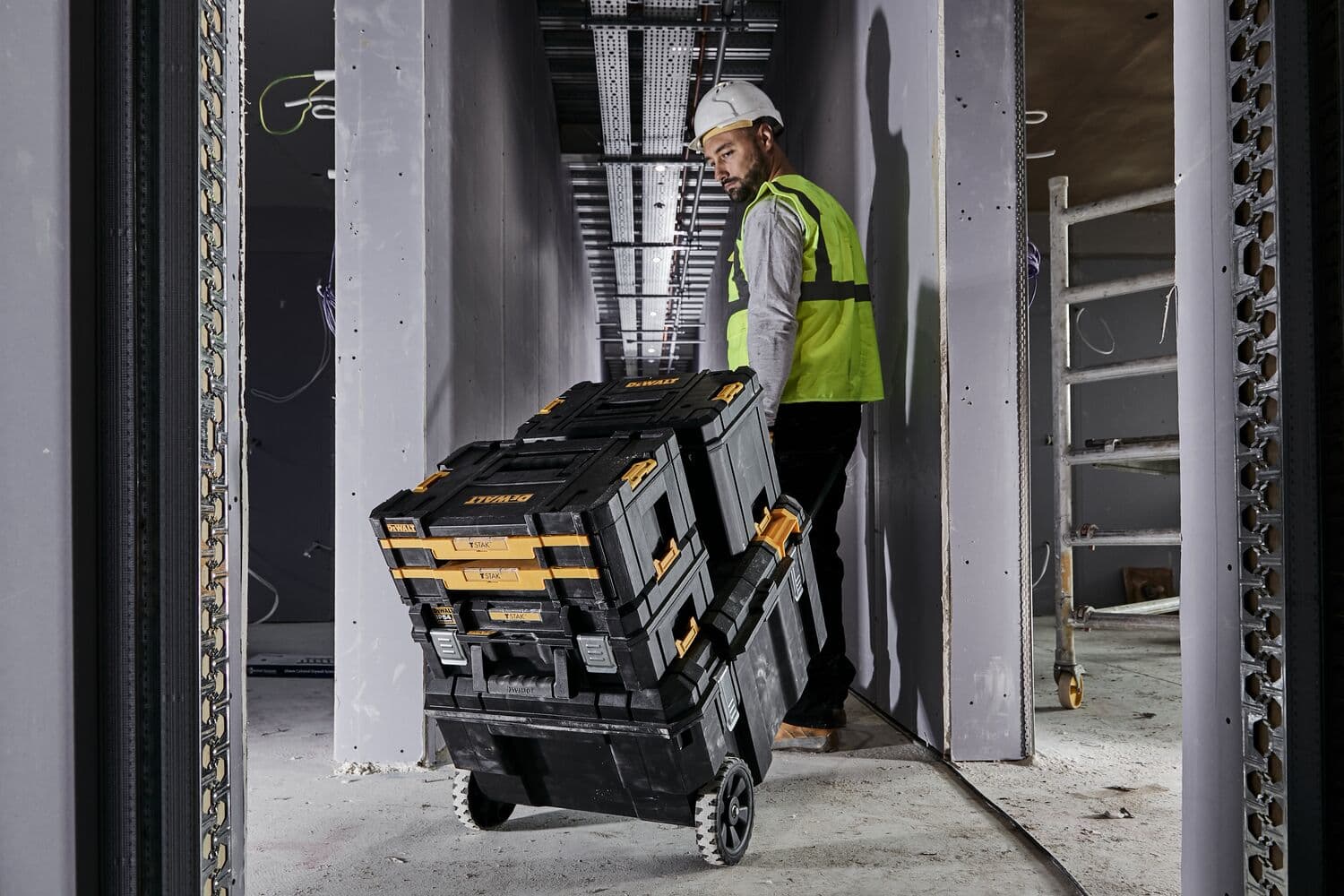 A person wearing a safety vest and hard hat pulls a stack of DEWALT toolboxes on wheels through a construction site hallway.