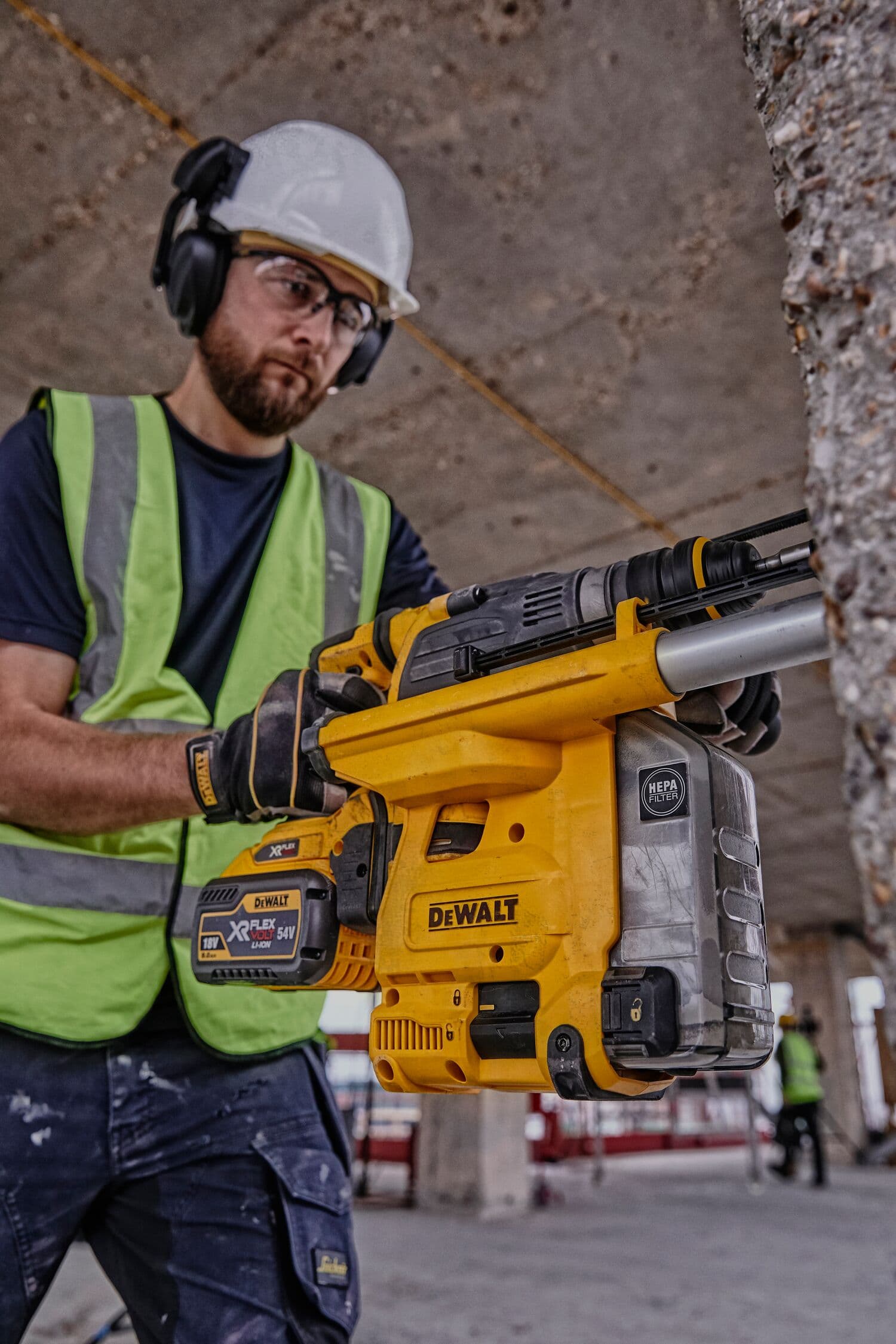 A person wearing safety gear and a high-visibility vest is holding a DEWALT DCH334X2 cordless rotary hammer drill with a battery, working at a construction site.