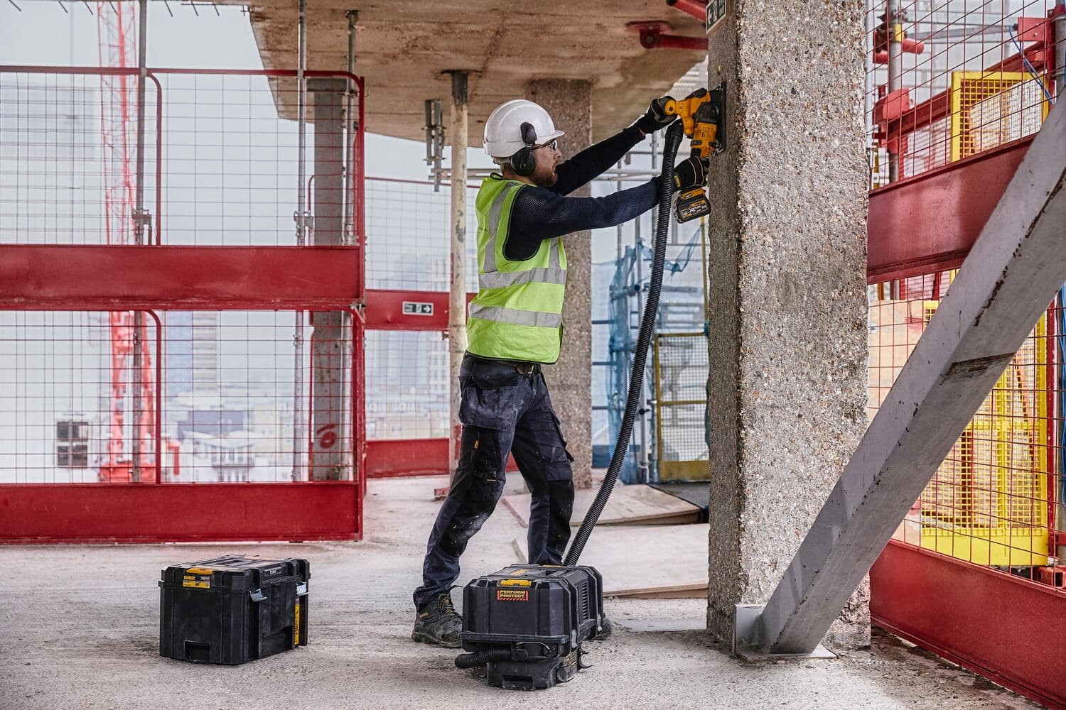 A construction worker wearing safety gear, including a helmet and high-visibility vest, uses a DEWALT power tool to cut into a concrete pillar at a construction site. DEWALT storage and equipment boxes are positioned on the ground nearby.