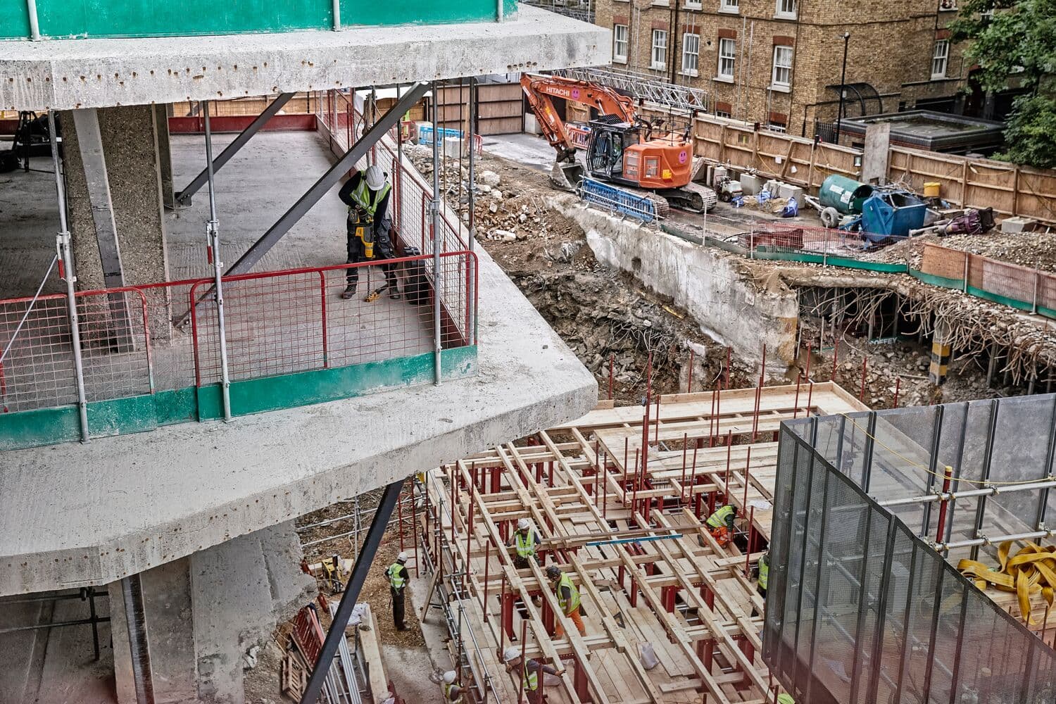 Construction site featuring an unfinished multi-story concrete structure. Workers wearing safety gear are present on different levels and in various areas, including one using a tool on the upper floor. Below, scaffolding and wooden planks are being assembled, and an excavator operates near the edge of the site.