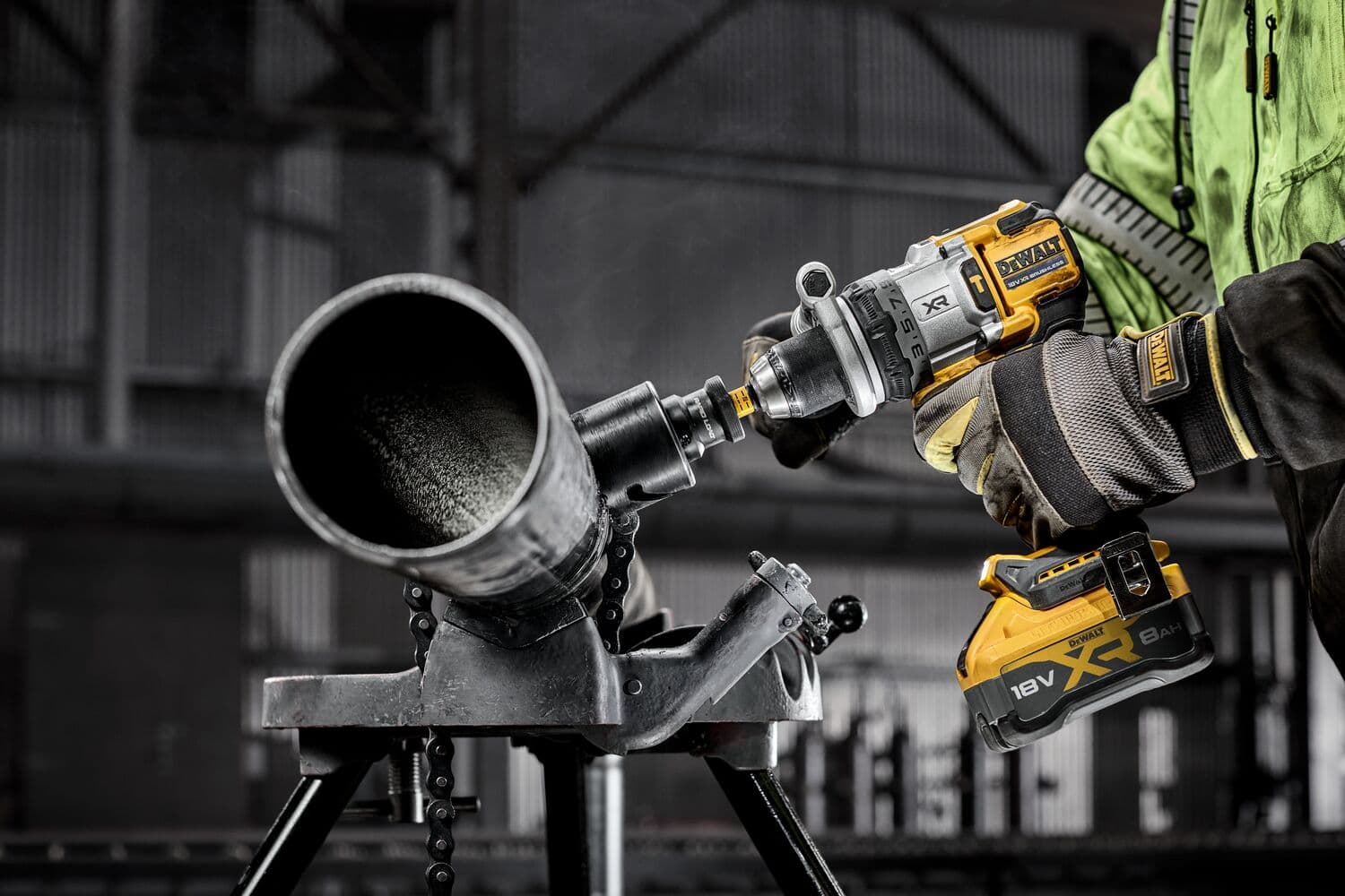 A person wearing gloves and a green jacket uses a DEWALT 18V XR cordless drill to cut into a large metal pipe mounted on a stand in an industrial setting.