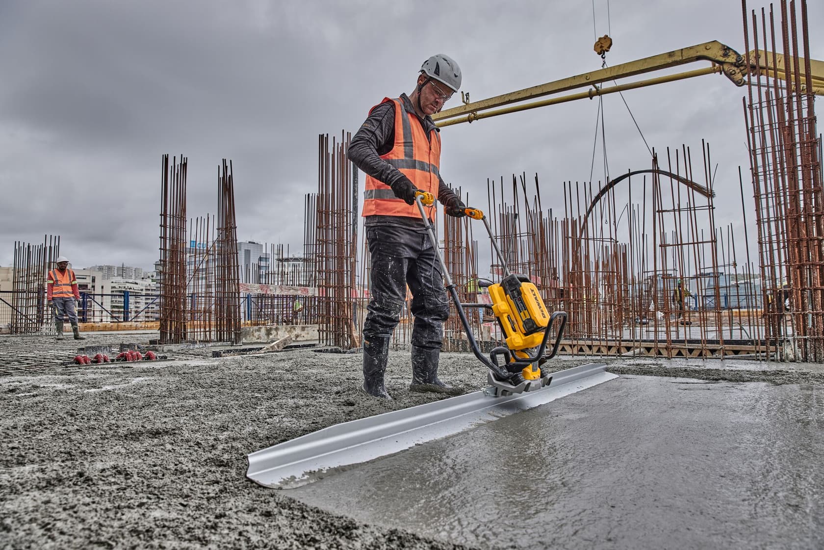 A construction worker wearing an orange safety vest and helmet operates a DEWALT power tool to smooth freshly poured concrete at a building site with exposed steel rods and construction equipment under a cloudy sky.