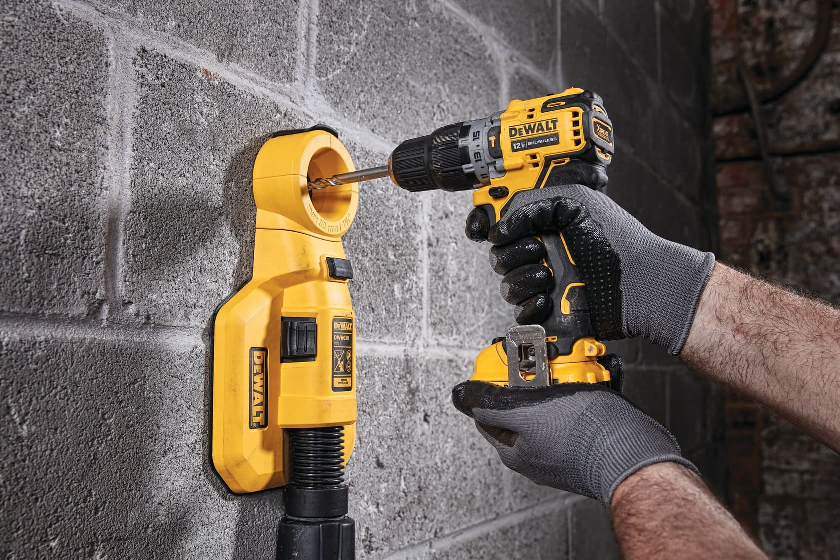 A person wearing gloves is using a DEWALT cordless drill to drill into a concrete wall, while a yellow DEWALT wall-mounted vacuum attachment is collecting dust next to the drill bit.