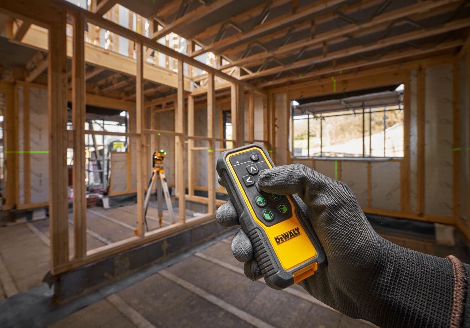 A gloved hand holds a DEWALT remote control in the foreground, with a DEWALT laser level on a tripod projecting green lines