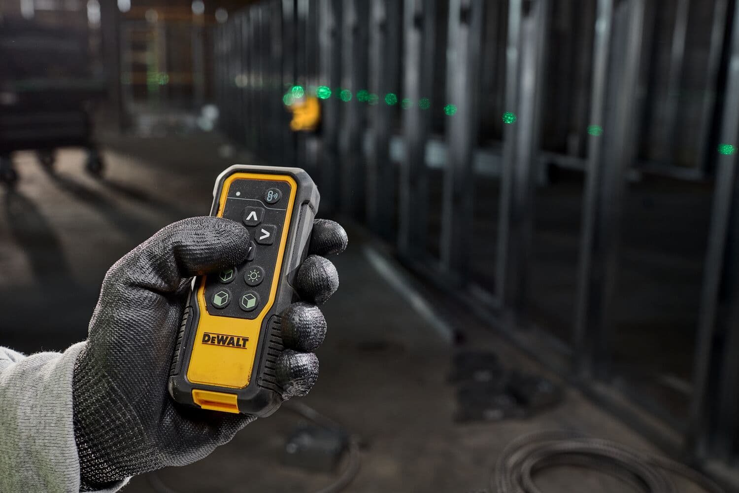 A gloved hand holding a DEWALT remote control device in an industrial setting, with a green laser line visible in the background.