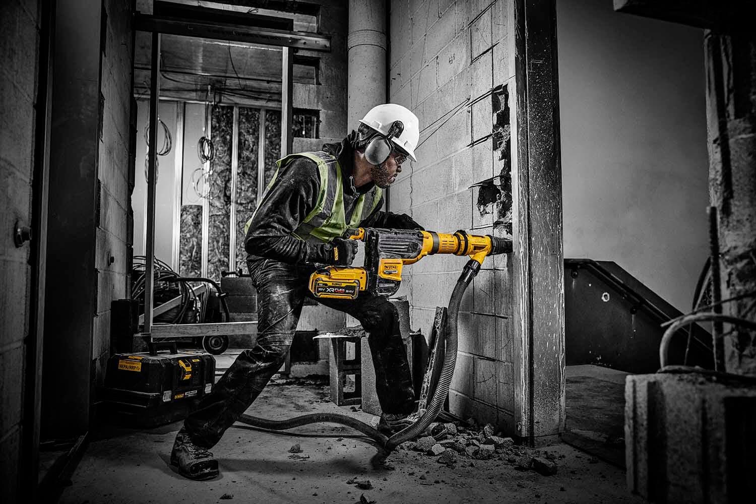 A construction worker wearing a hard hat and safety vest uses a DEWALT DCH775 rotary hammer drill to break through a concrete wall inside a building under renovation. Debris is scattered on the floor.
