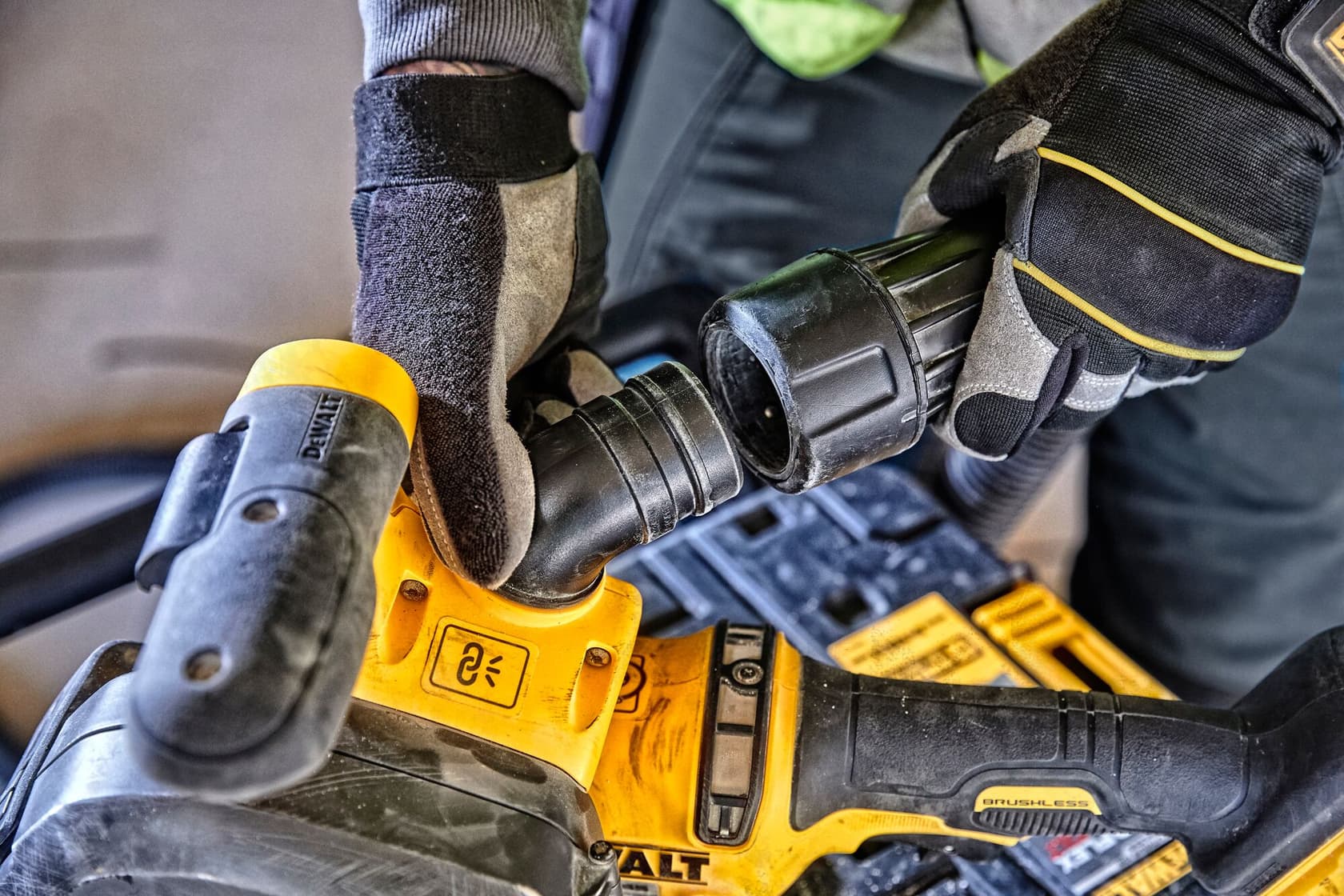 Close-up view of gloved hands connecting a hose to a DEWALT power tool, specifically showing the attachment process and the tool's handle and control buttons.