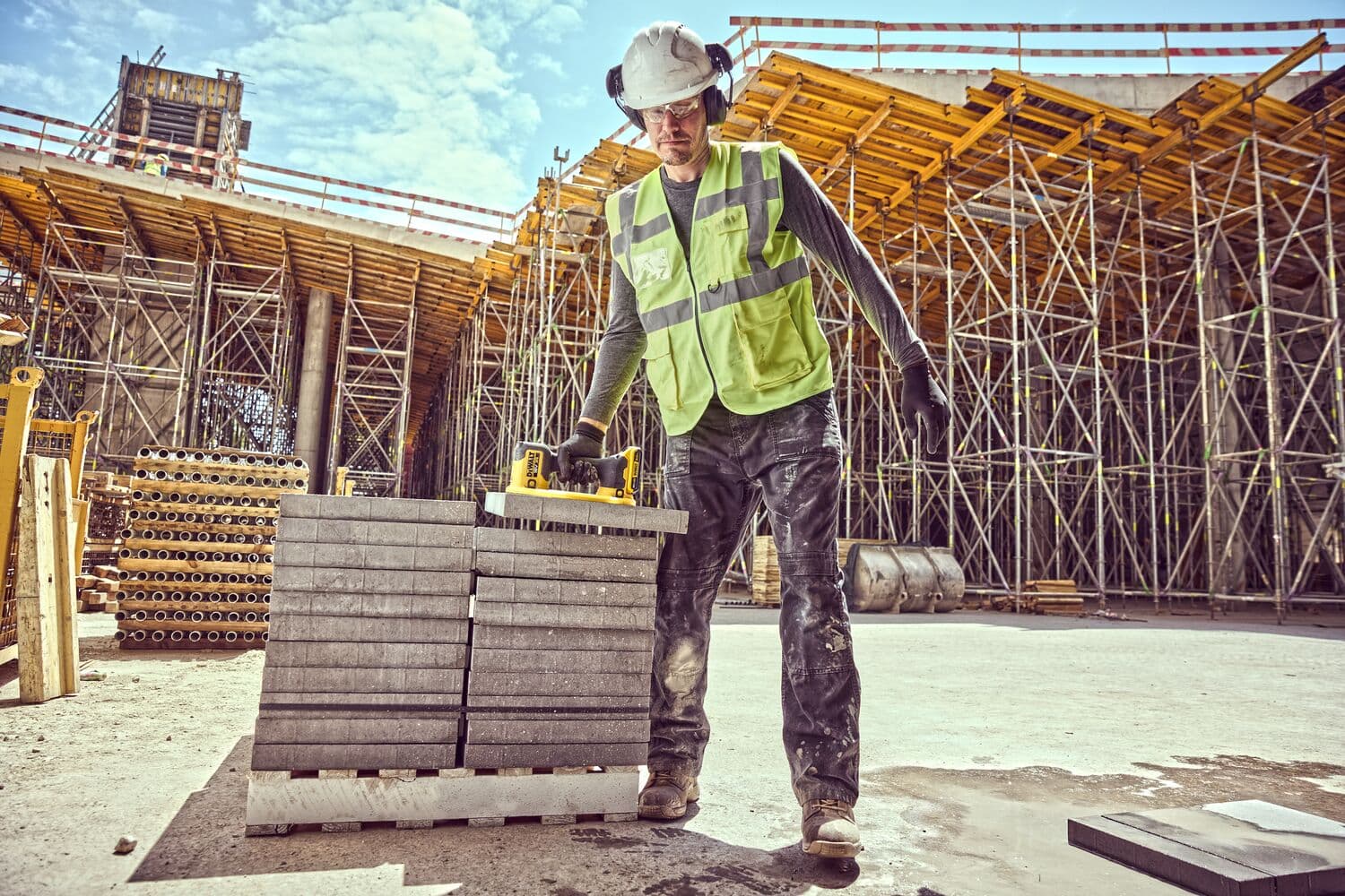 A construction worker wearing a helmet, ear protection, gloves, and a high-visibility vest uses a DEWALT DCE590 tool to lift