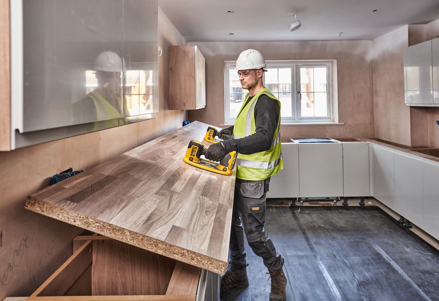 A construction worker wearing a safety helmet and high-visibility vest is installing a wooden countertop in a kitchen using