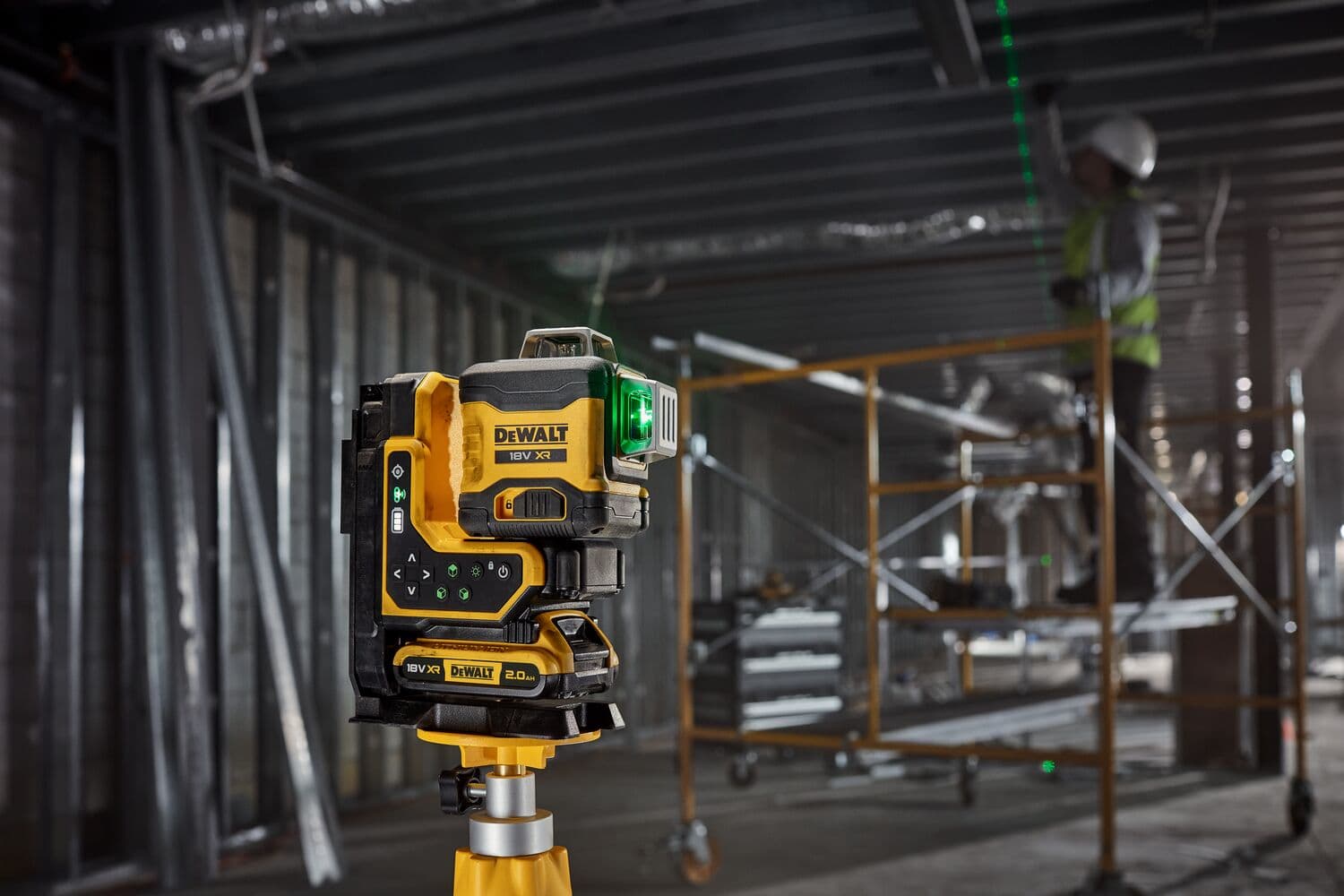 A DEWALT laser level device, model DCLE34035D1, mounted on a tripod in a construction site with scaffolding and a worker in