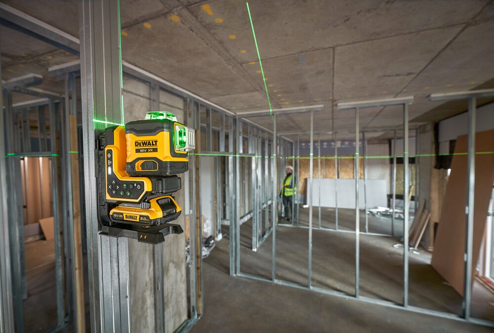 A DEWALT green line laser tool mounted on a metal frame inside a building under construction. The laser projects green lines