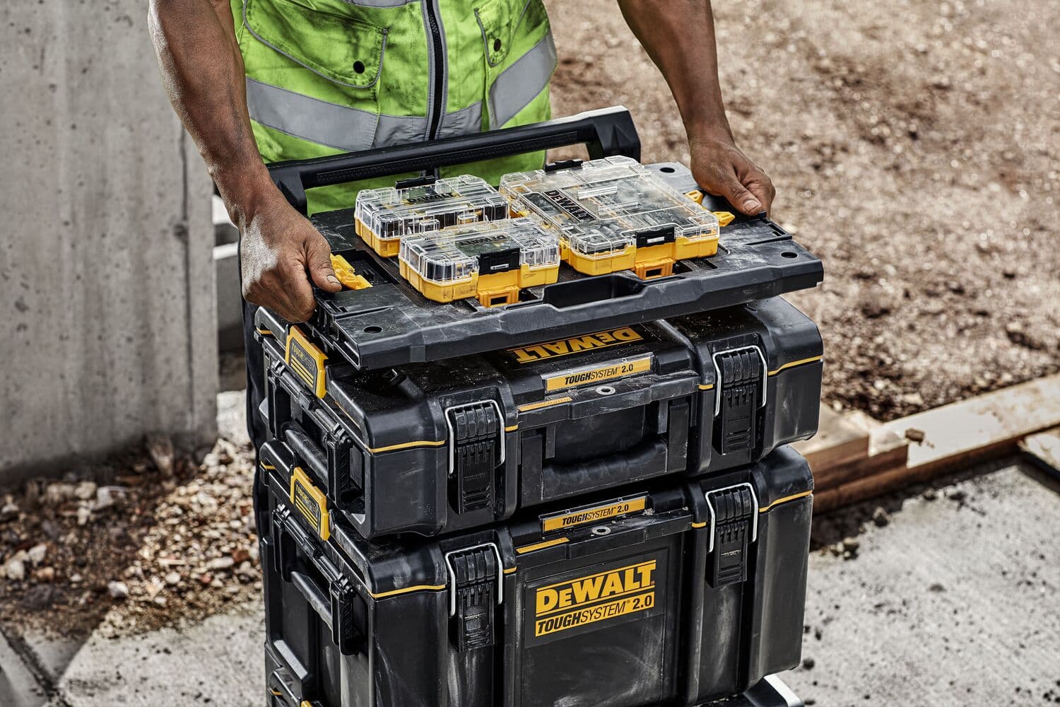 A person in a high-visibility vest stacks several black DEWALT ToughSystem 2.0 toolboxes and organizers outdoors on a worksite.