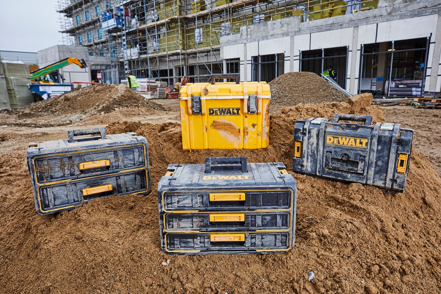 Four DEWALT toolboxes on a construction site dirt mound, with a building and workers in the background.