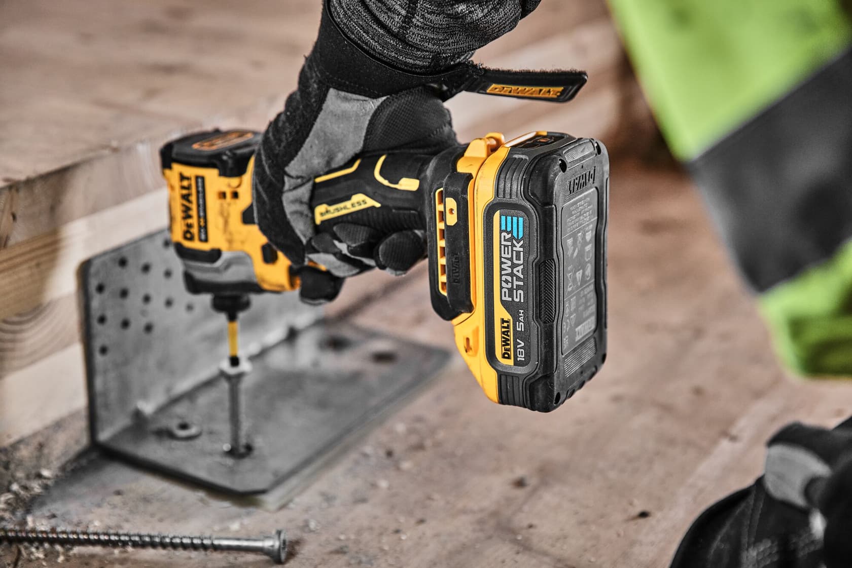 A person wearing protective gloves uses a yellow and black DEWALT cordless power drill with a POWERSTACK battery to drive a screw into a metal bracket attached to a wooden beam at a construction site.