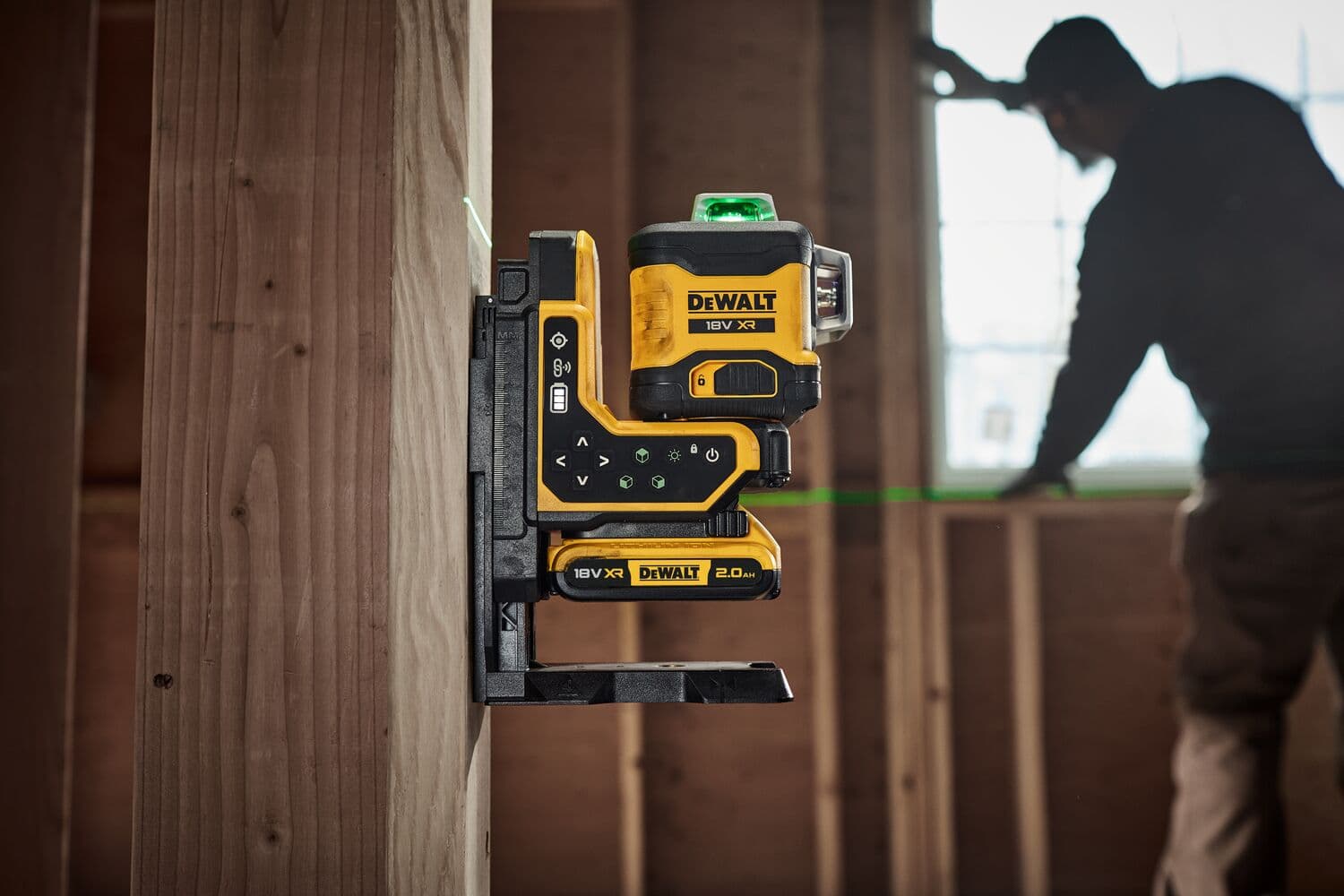 A DEWALT 18V XR green laser level tool (model DCLE34035D1) is mounted on a wooden stud, projecting a green laser line across
