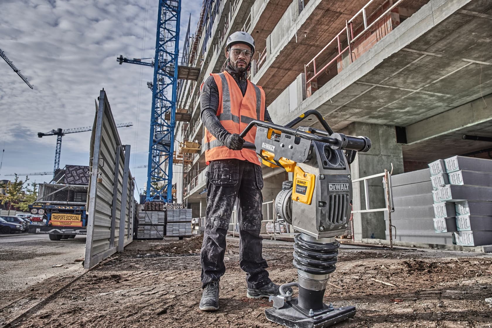 Worker using a DEWALT compactor at a building site, wearing safety gear. Construction materials are visible nearby.