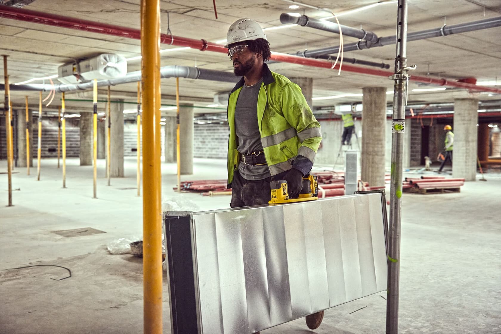 A construction worker wearing a high-visibility jacket and hard hat is carrying a large metal panel using a DEWALT tool