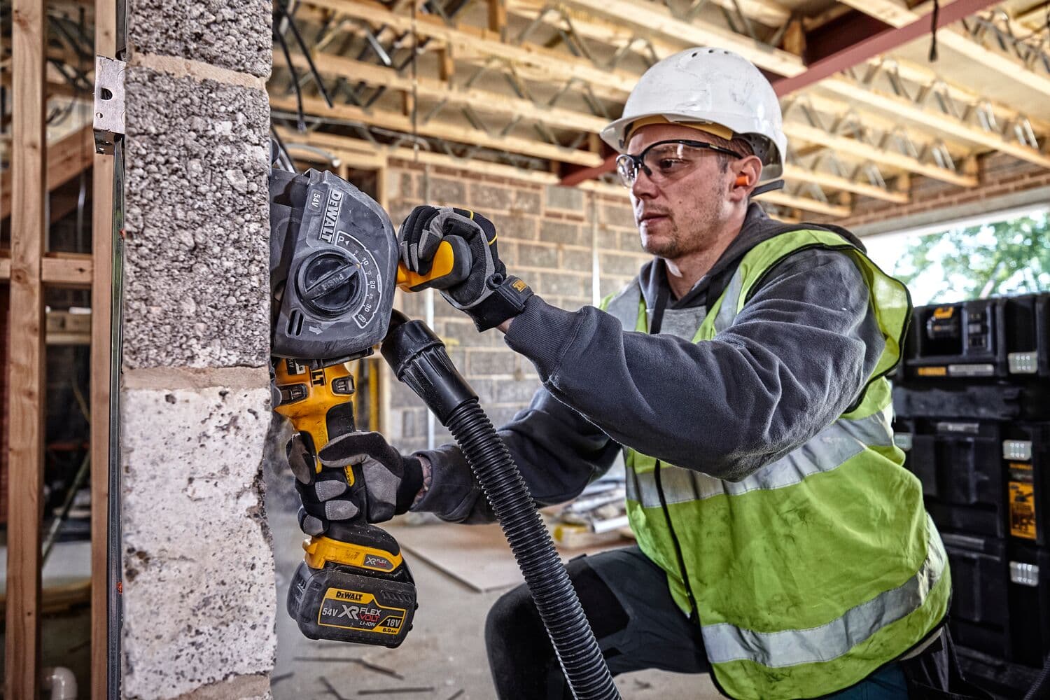Person wearing a safety helmet and high-visibility vest using a DEWALT cordless wall chaser tool on a concrete block wall at a construction site. The tool is equipped with a dust extraction hose. DEWALT storage boxes are visible in the background.