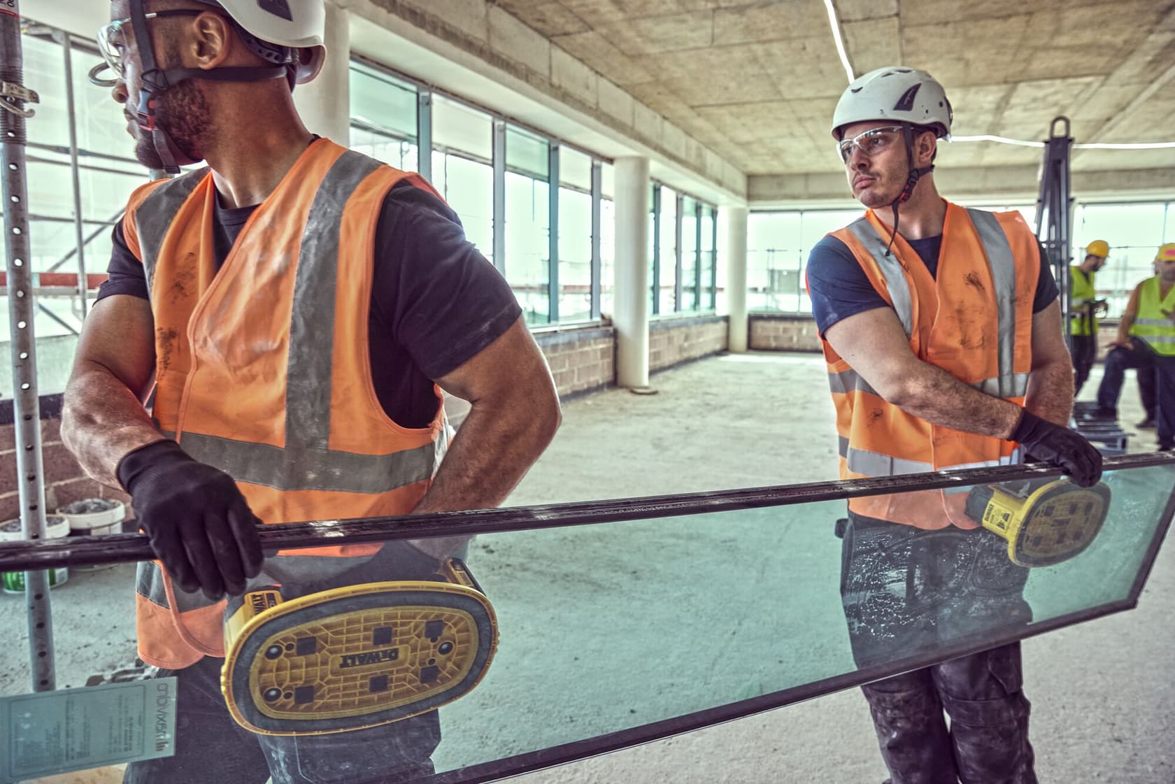 Two construction workers wearing safety helmets and orange vests use DEWALT DCE590 suction tools to carry a large glass